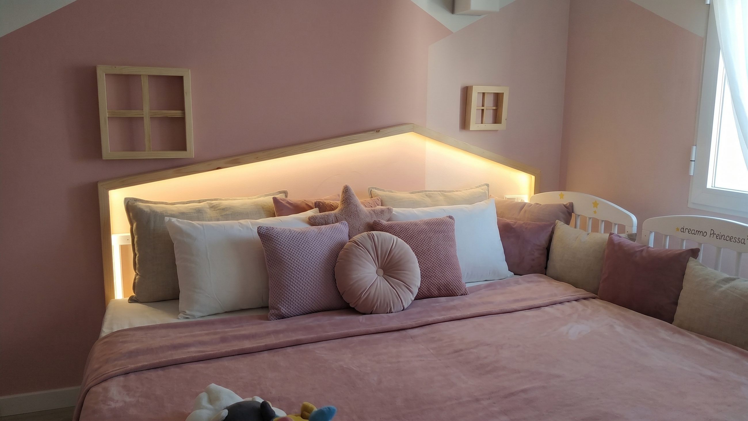 Cabecero de madera en forma de casa con luz indirecta sobre pared rosa